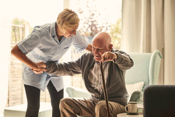 Smiling senior man with female healthcare worker. Home carer supporting old man to stand up from the chair at care home.